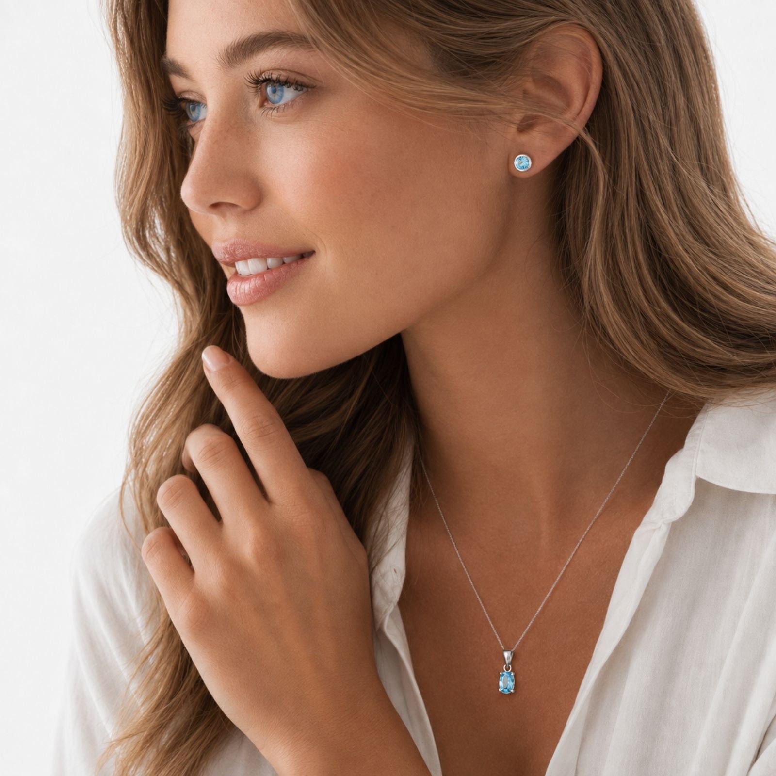 Woman with long brown hair wearing a white shirt. She has a blue stone earring and a matching blue pendant necklace. She is looking to the side and gently touching her face with her hand.