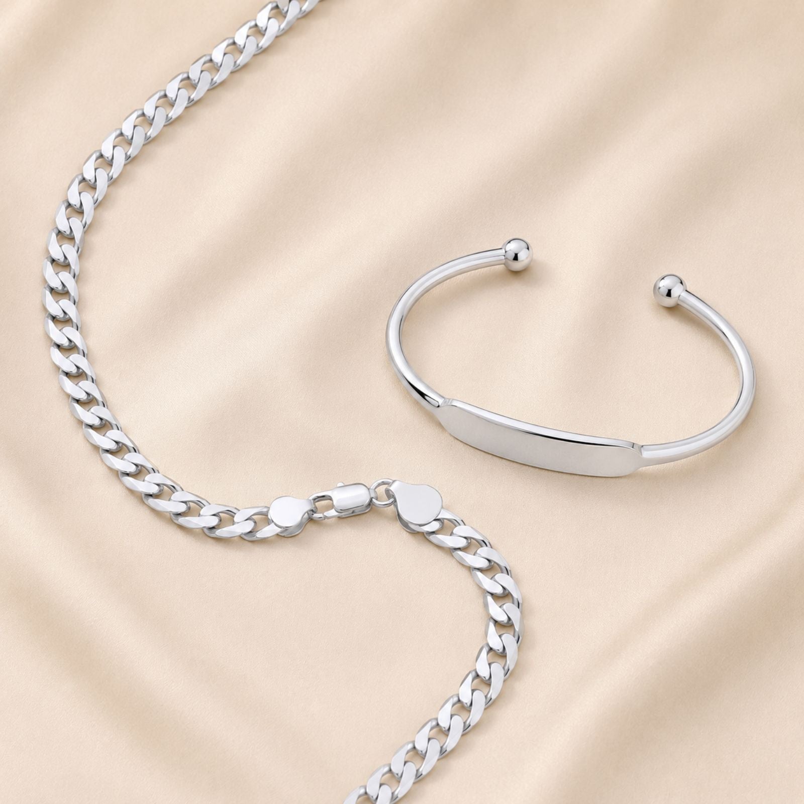 A man with short curly dark hair and a beard wears a dark sweater, silver chain necklace, bracelet, and ring, standing against a neutral background, looking slightly to the side with a slight smile.