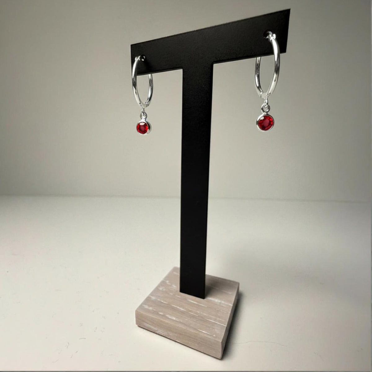 Sterling silver hoop earrings with simulated garnet charms displayed on a black T-stand with light wood base.