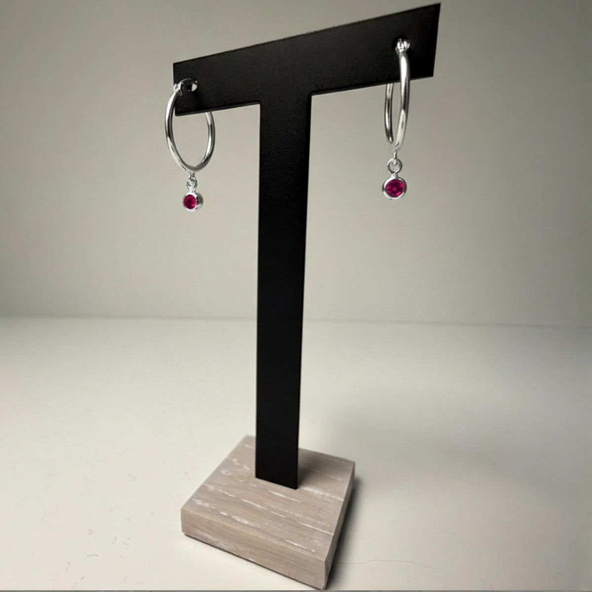 Sterling silver hoop earrings with simulated ruby charms shown on a black T-stand featuring wood accents.