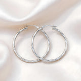A pair of polished silver hoop earrings with a twisted rope texture, displayed on a draped white satin fabric background.