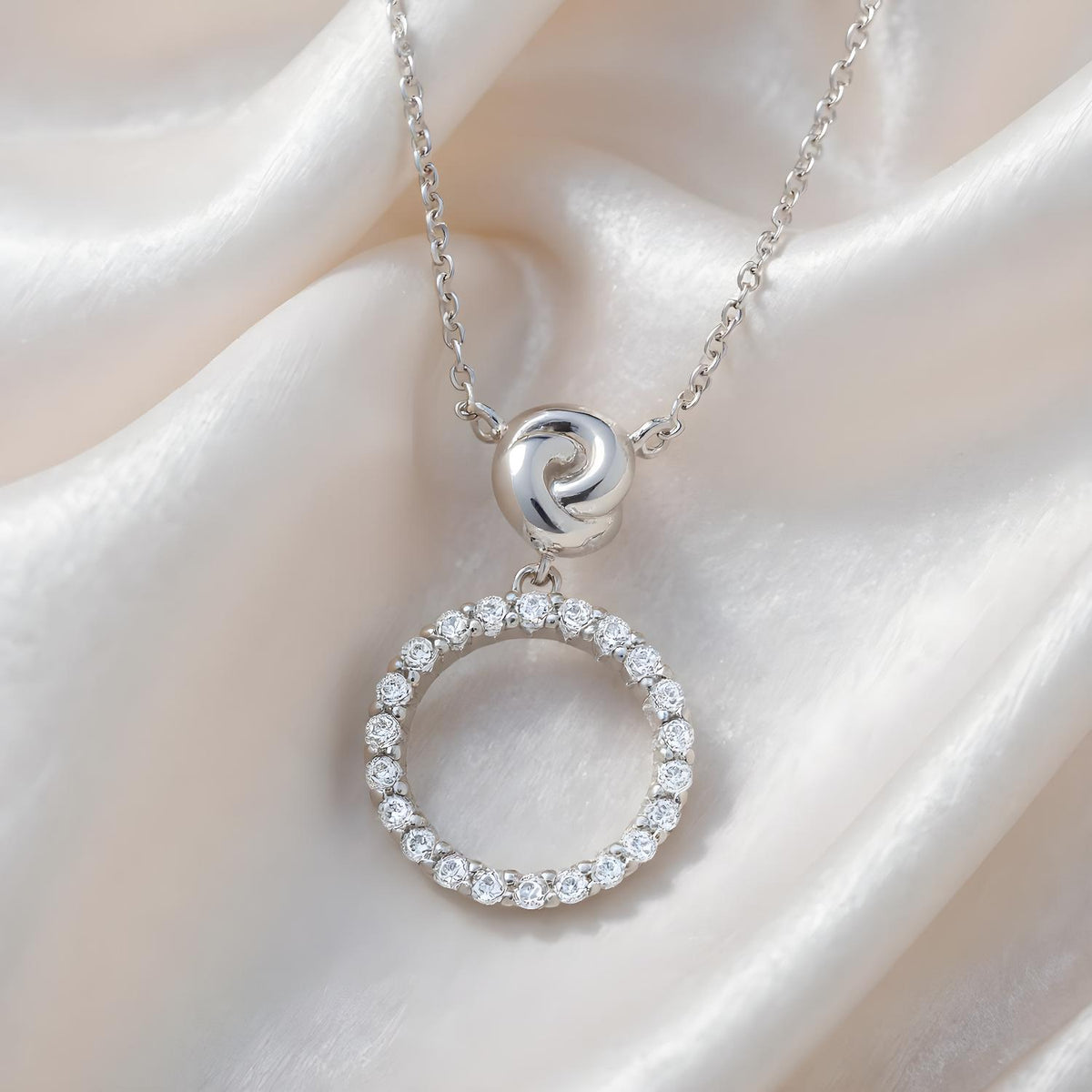 Sterling silver infinity love knot circle necklace with a cubic zirconia pendant, displayed on white satin.