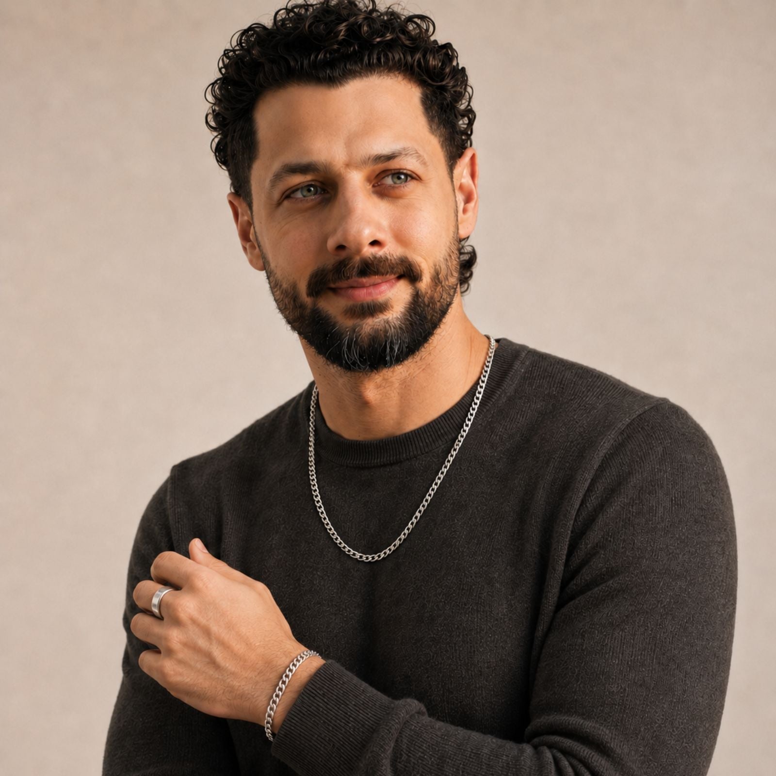 A man with short curly dark hair and a beard wears a dark sweater, silver chain necklace, bracelet, and ring, standing against a neutral background, looking slightly to the side with a slight smile.