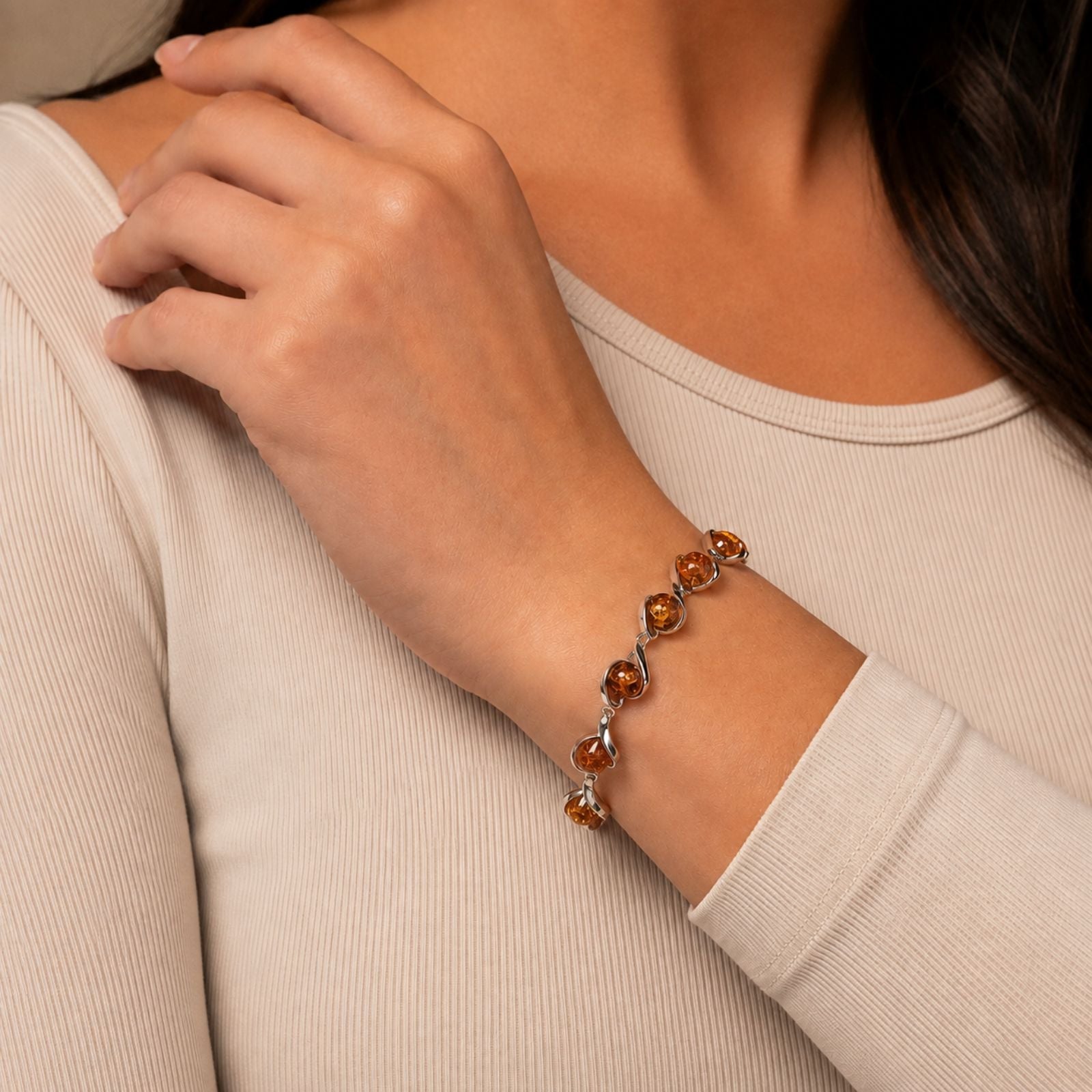 A person wearing a long-sleeve beige top displays a gold bracelet with multiple amber-colored gemstones on their wrist. Their hand rests gently near their collarbone.