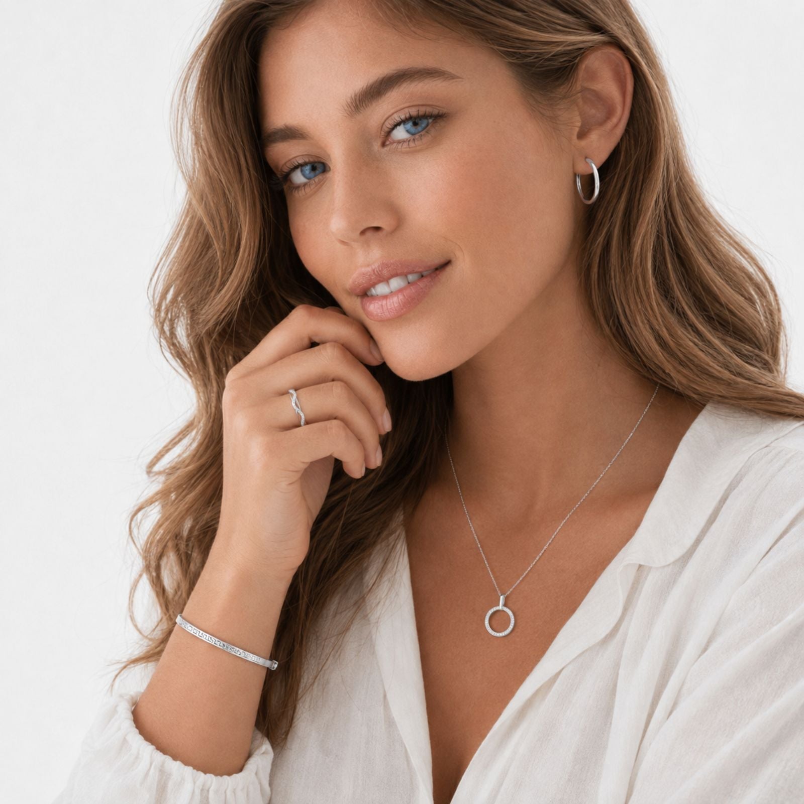 Woman with wavy brown hair wears a white blouse, silver hoop earrings, a circular pendant necklace, a thin bracelet, and a ring. She is looking at the camera with a slight smile and hand near her face.