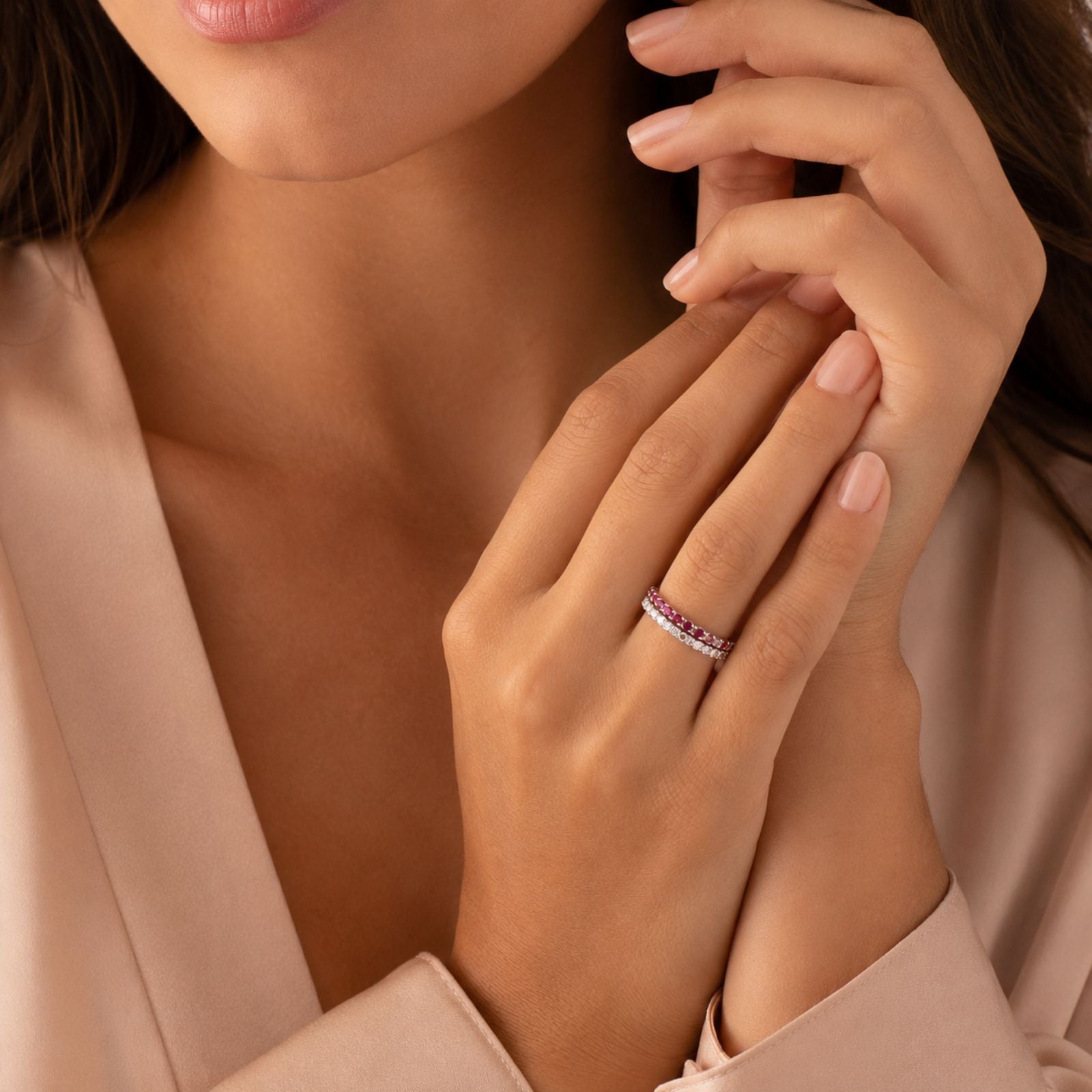 A woman wearing a light beige blouse displays her hands, one of which has a ring with pink and white stones. Her nails are manicured with a pale polish. Her face is partially visible.