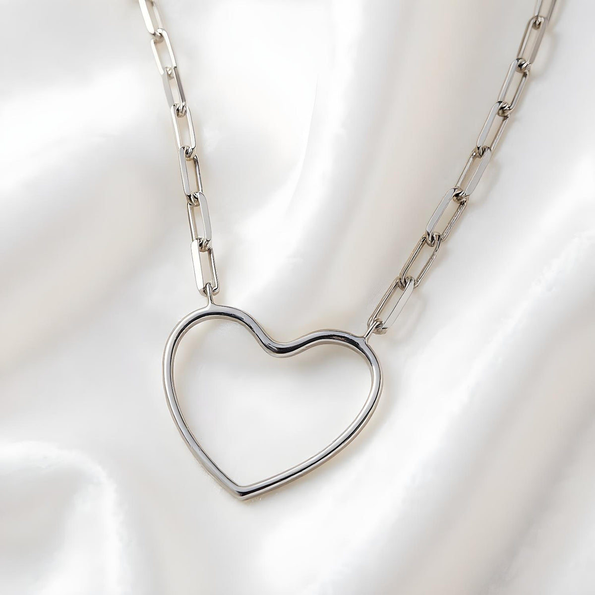 A sterling silver paperclip chain necklace with an open heart pendant lies on smooth white satin fabric.