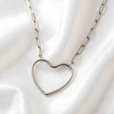 A sterling silver paperclip chain necklace with an open heart pendant lies on smooth white satin fabric.