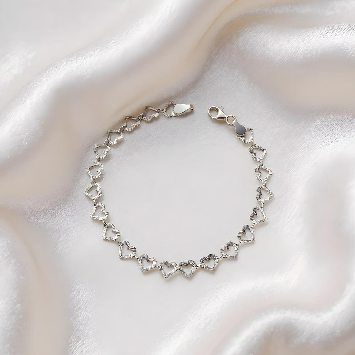 A sterling silver bracelet with diamond-cut heart links is displayed on white satin fabric.