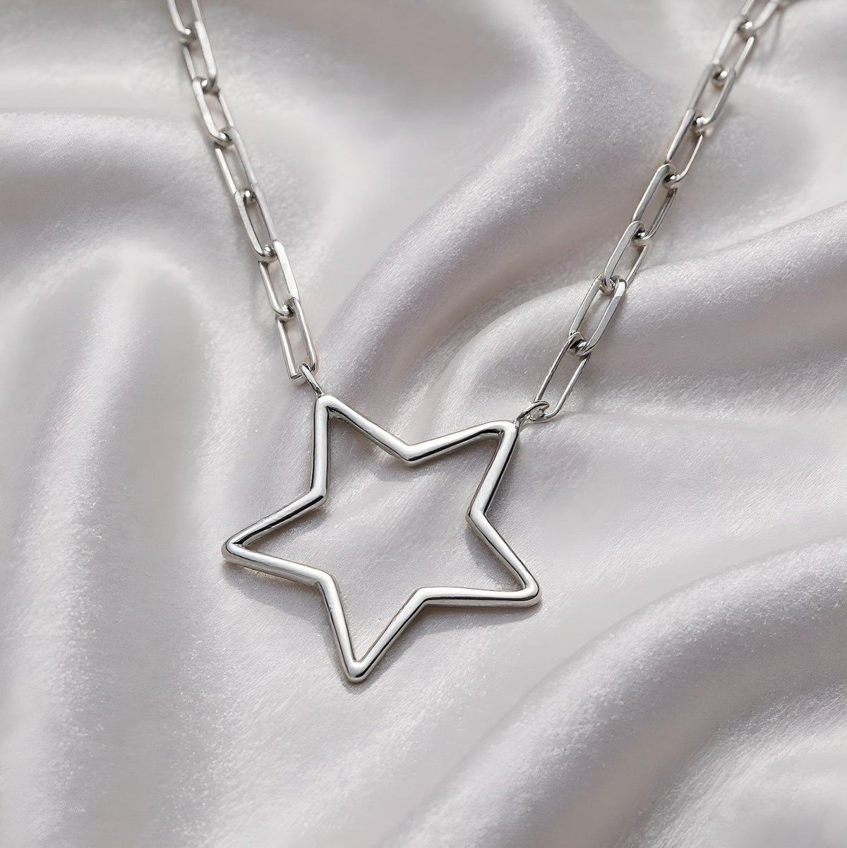 A Sterling silver paperclip chain necklace with an open star pendant rests on smooth light-colored satin fabric.