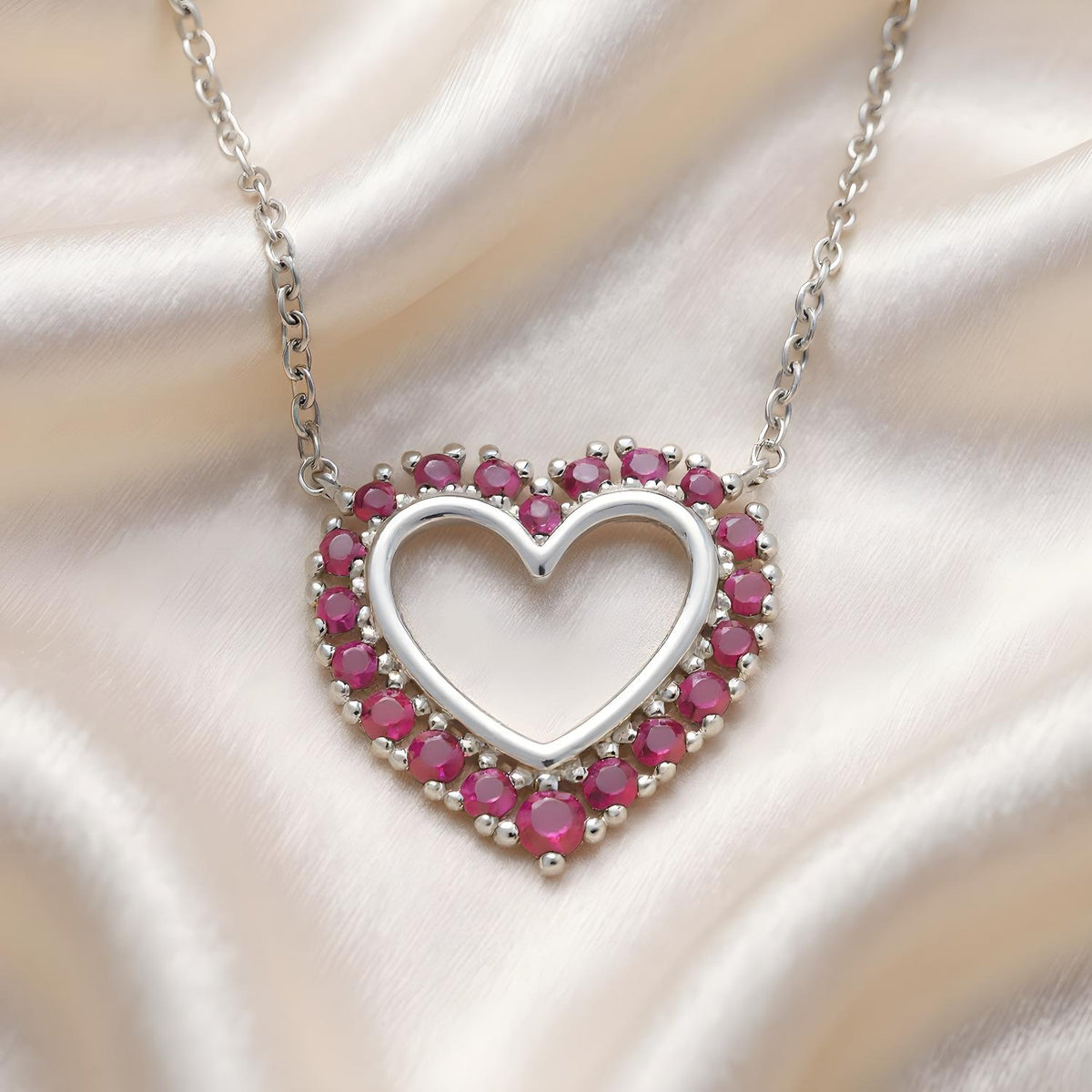 Silversmiths Sterling Silver necklace with a simulated ruby open heart pendant, shown on soft satin fabric.