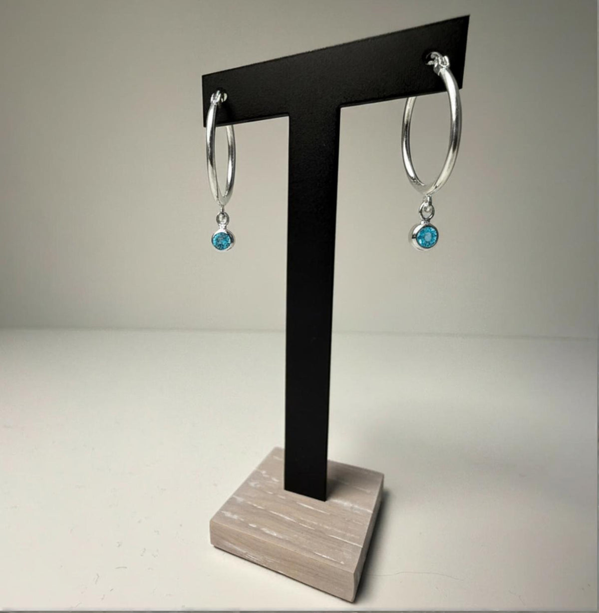 Sterling silver hoop earrings with simulated London topaz charms displayed on a black T-stand with a wood base.
