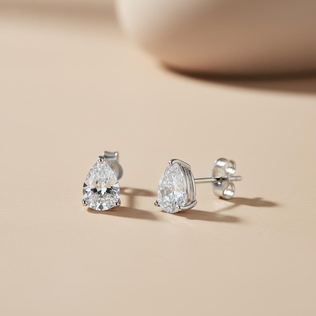 A pair of Silversmiths 7x5mm sterling silver teardrop CZ stud earrings, one standing upright on a beige surface.