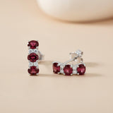 Sterling silver drop stud earrings with three simulated rubies and clear stones on a beige background.