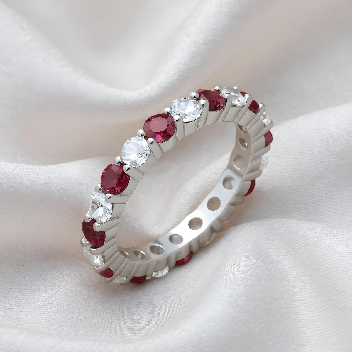 A sterling silver eternity ring with simulated rubies and CZ stones is displayed on white satin fabric.