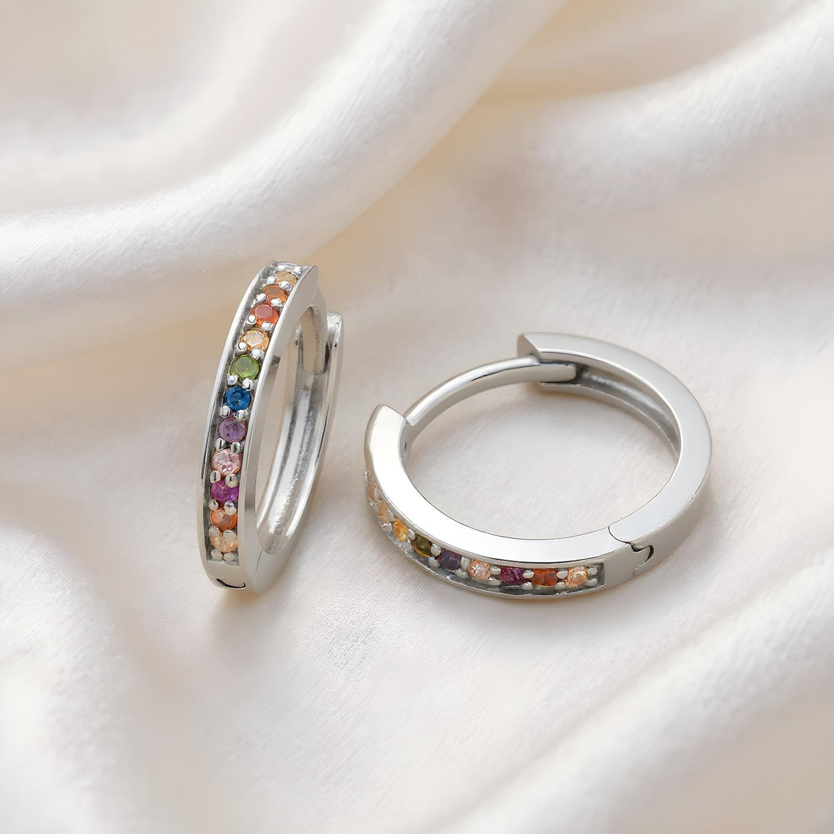 Sterling silver huggie hoops with rainbow cubic zirconia rest on smooth white silky fabric.