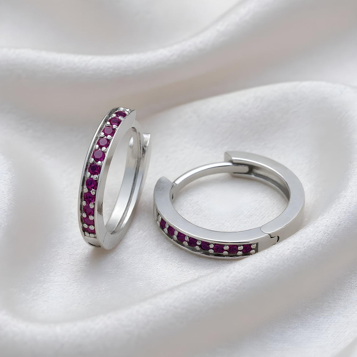 Sterling silver huggie hoop earrings with simulated amethyst rest on a white satin fabric background.