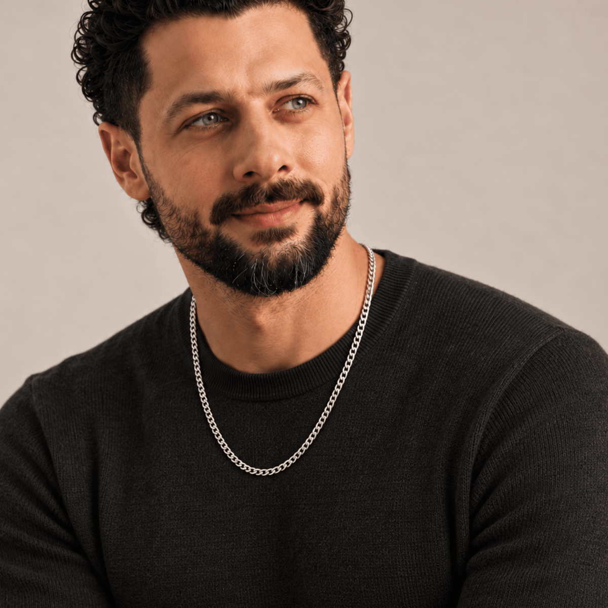 Man with curly dark hair and beard wears a black sweater and silver chain necklace, looking to the side.