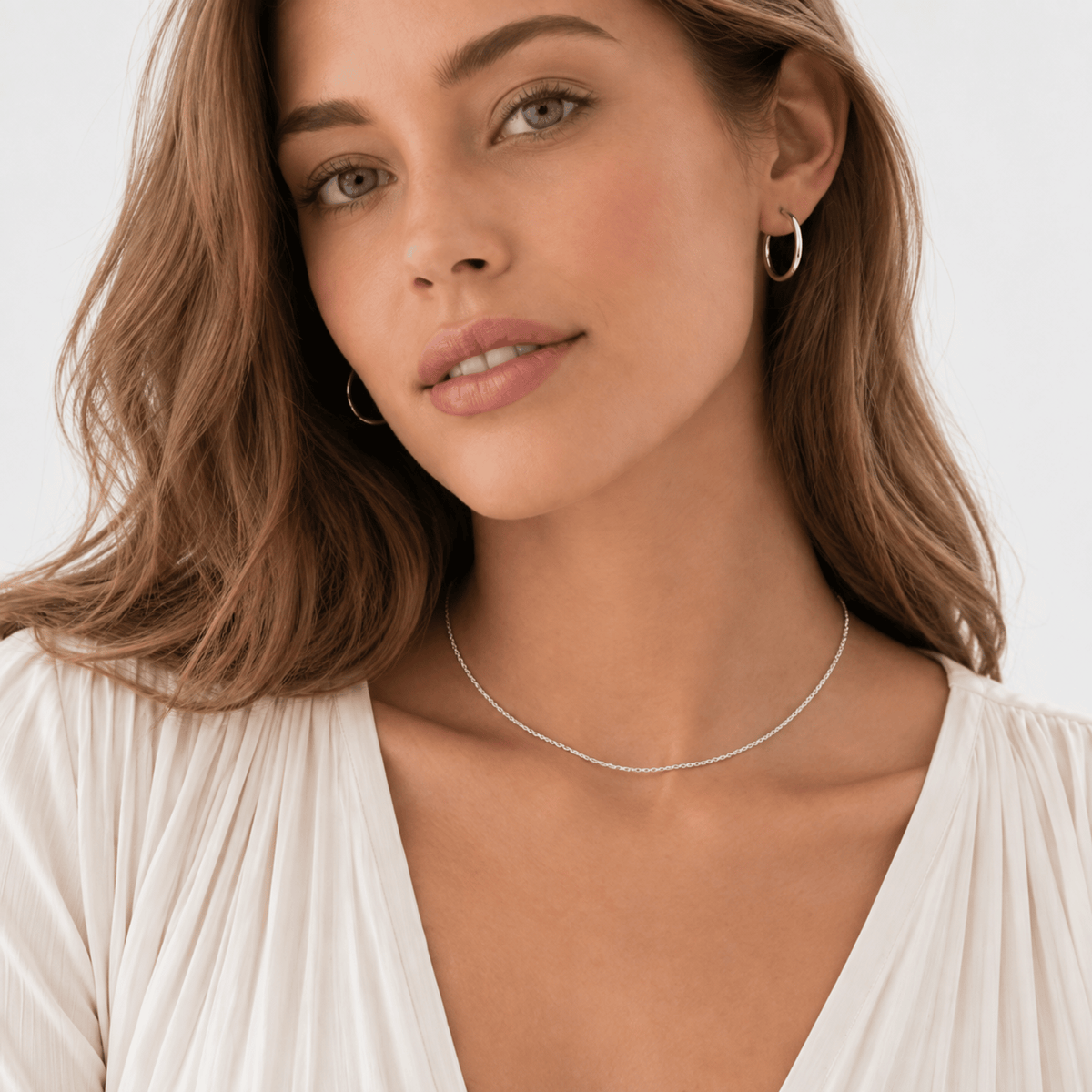 A woman with wavy brown hair wears hoop earrings, a thin necklace, and a white pleated top while looking at the camera.