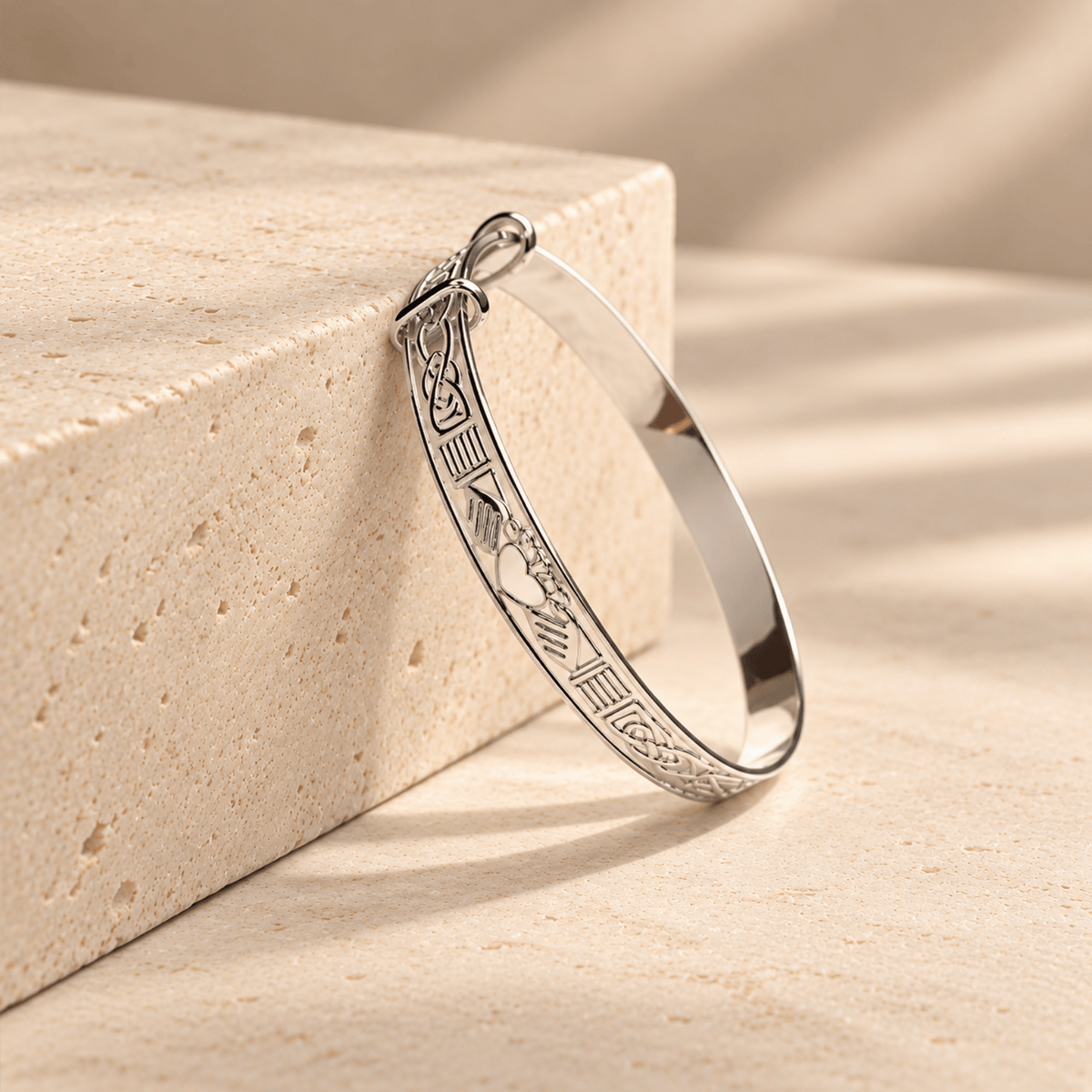 A sterling silver expanding baby bangle with a Claddagh motif rests on a beige block in natural sunlight.