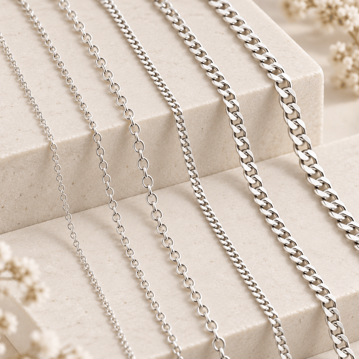 Six silver chains with different link designs rest on beige stone steps with dried flowers in the background.