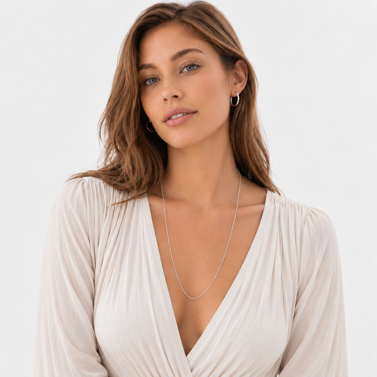 A woman with wavy brown hair is shown wearing a cream blouse and silver hoop earrings against a plain background.