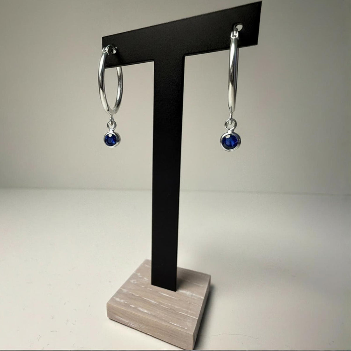 Sterling silver simulated sapphire charm hoop earrings displayed on a black T-stand with a wood base.