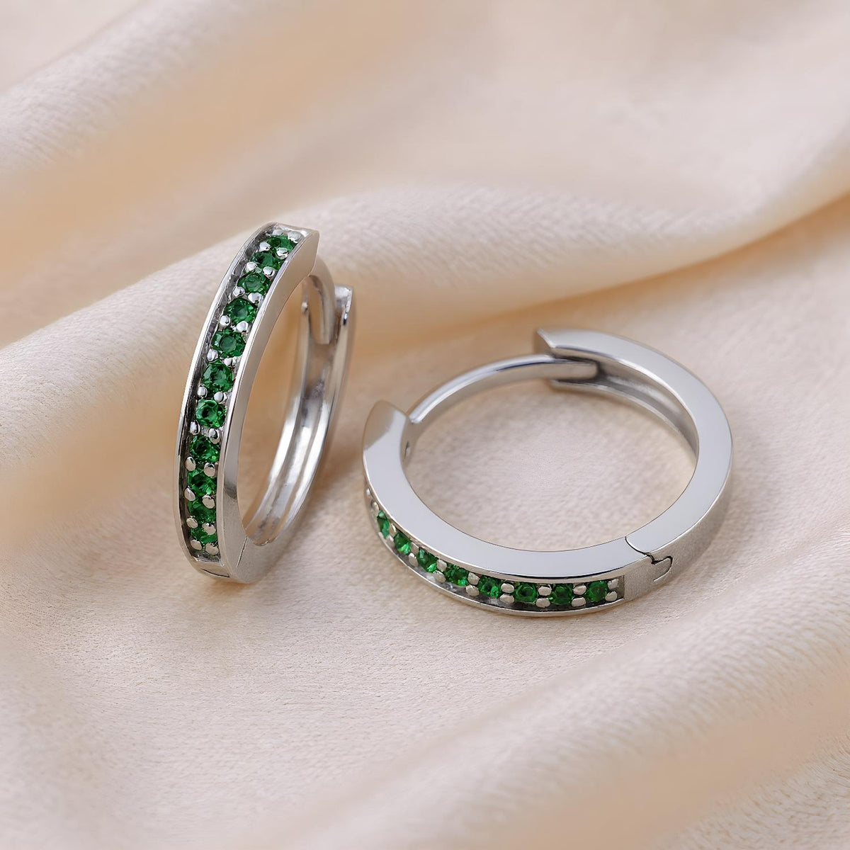 Silversmiths sterling silver huggie hoop earrings with simulated emeralds on a cream fabric background.