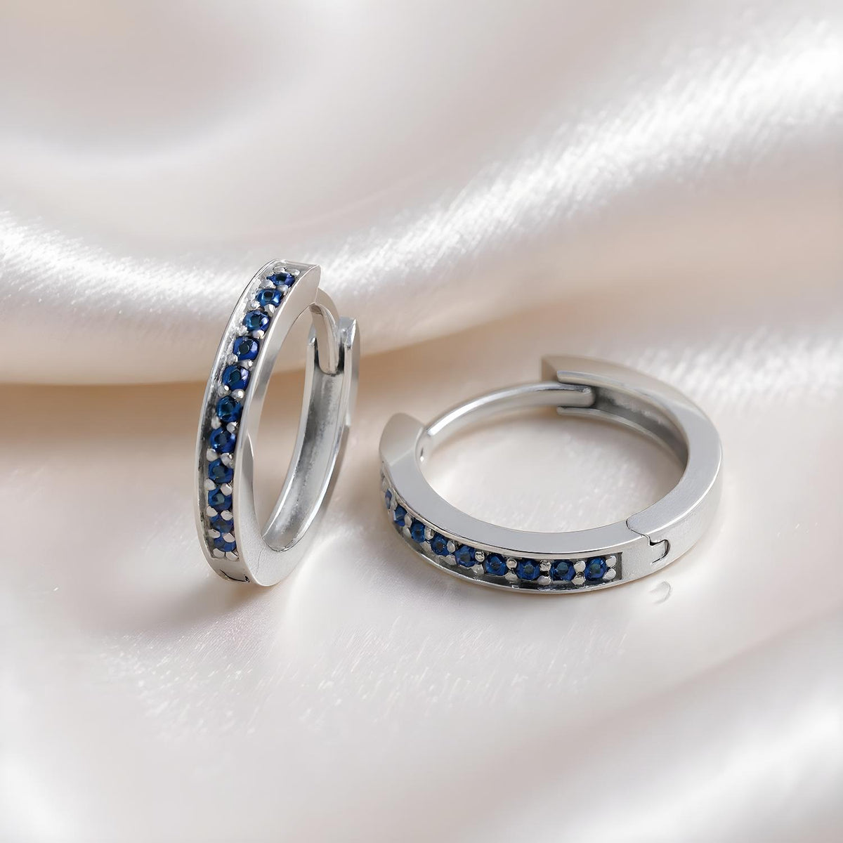 Sterling silver huggie hoop earrings with simulated sapphires, shown on smooth white satin fabric.