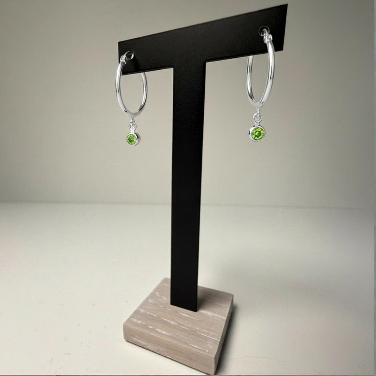 Sterling silver hoop earrings with simulated peridot charms displayed on a black T-stand with a wooden base.