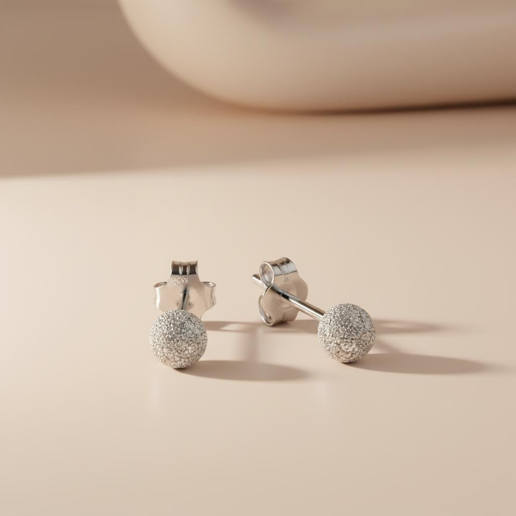 Silversmiths Sterling Silver 4mm frosted ball stud earrings rest on a smooth beige surface in soft lighting.