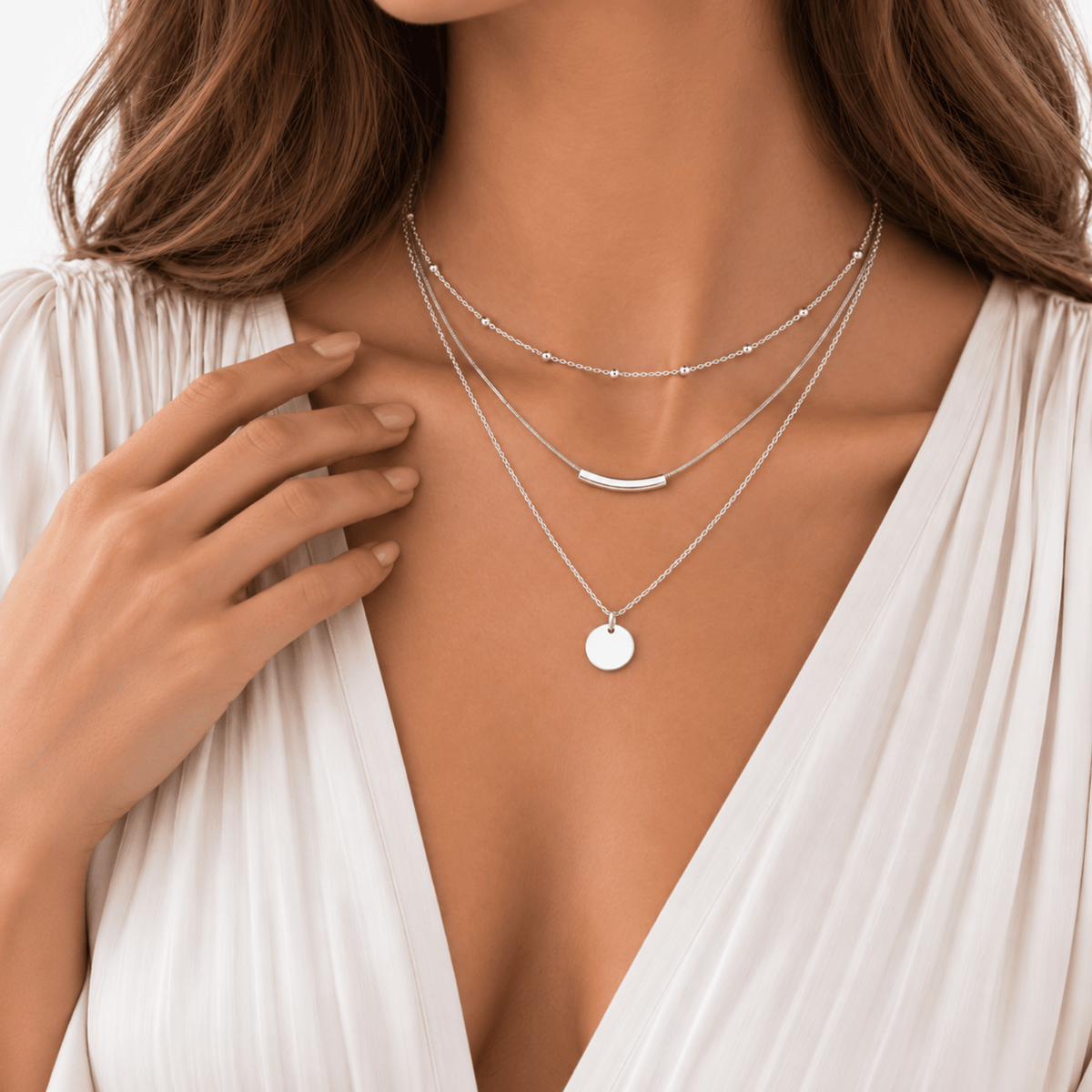 A woman wears three layered silver necklaces with a white v-neck blouse, her hand resting by her collarbone.