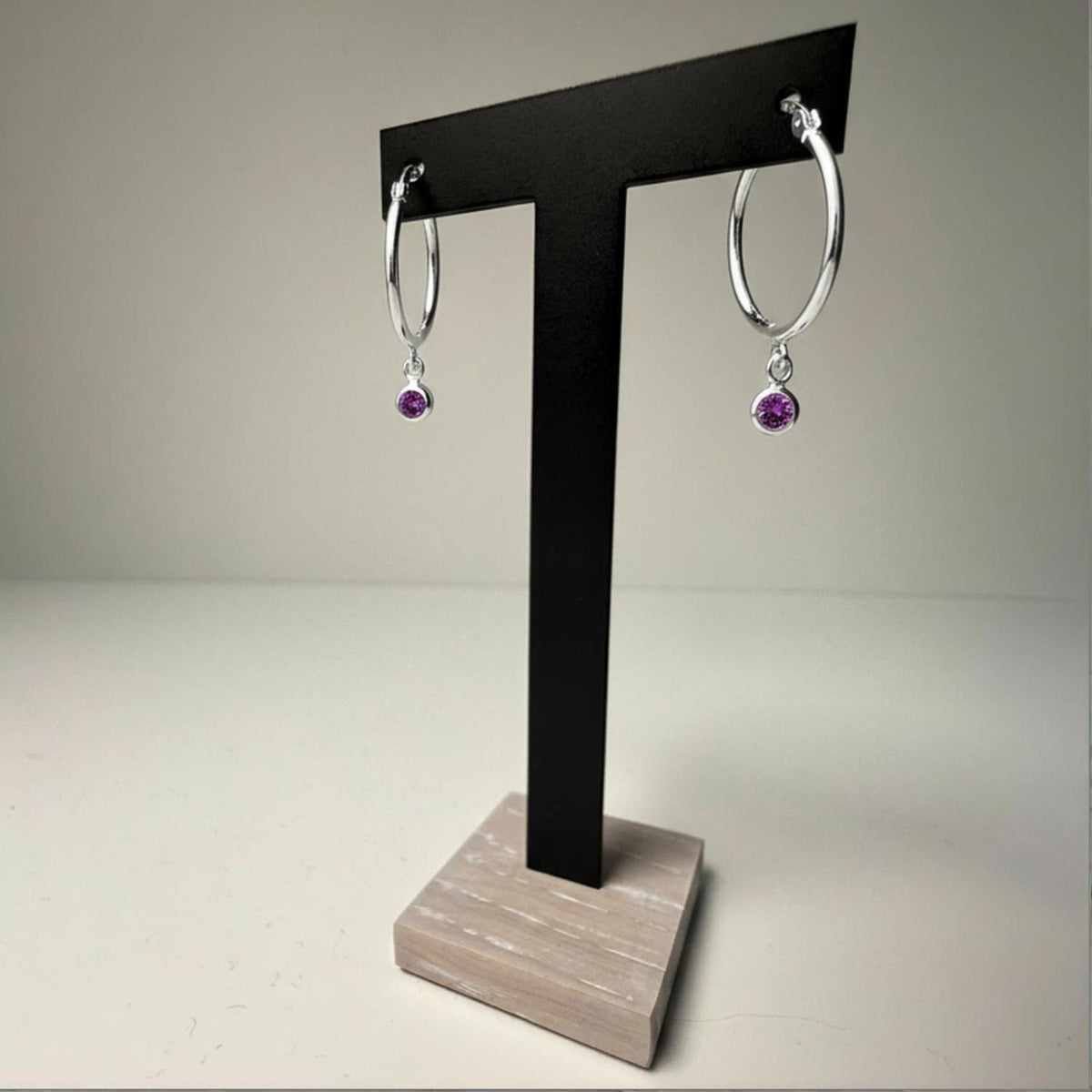 Silversmiths sterling silver hoop earrings with simulated amethyst charms on a black stand with a wooden base.