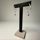 Silversmiths sterling silver hoop earrings with simulated amethyst charms on a black stand with a wooden base.