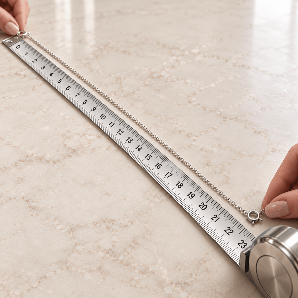 Hands hold a silver chain stretched along a metal ruler on a marble surface for measurement.