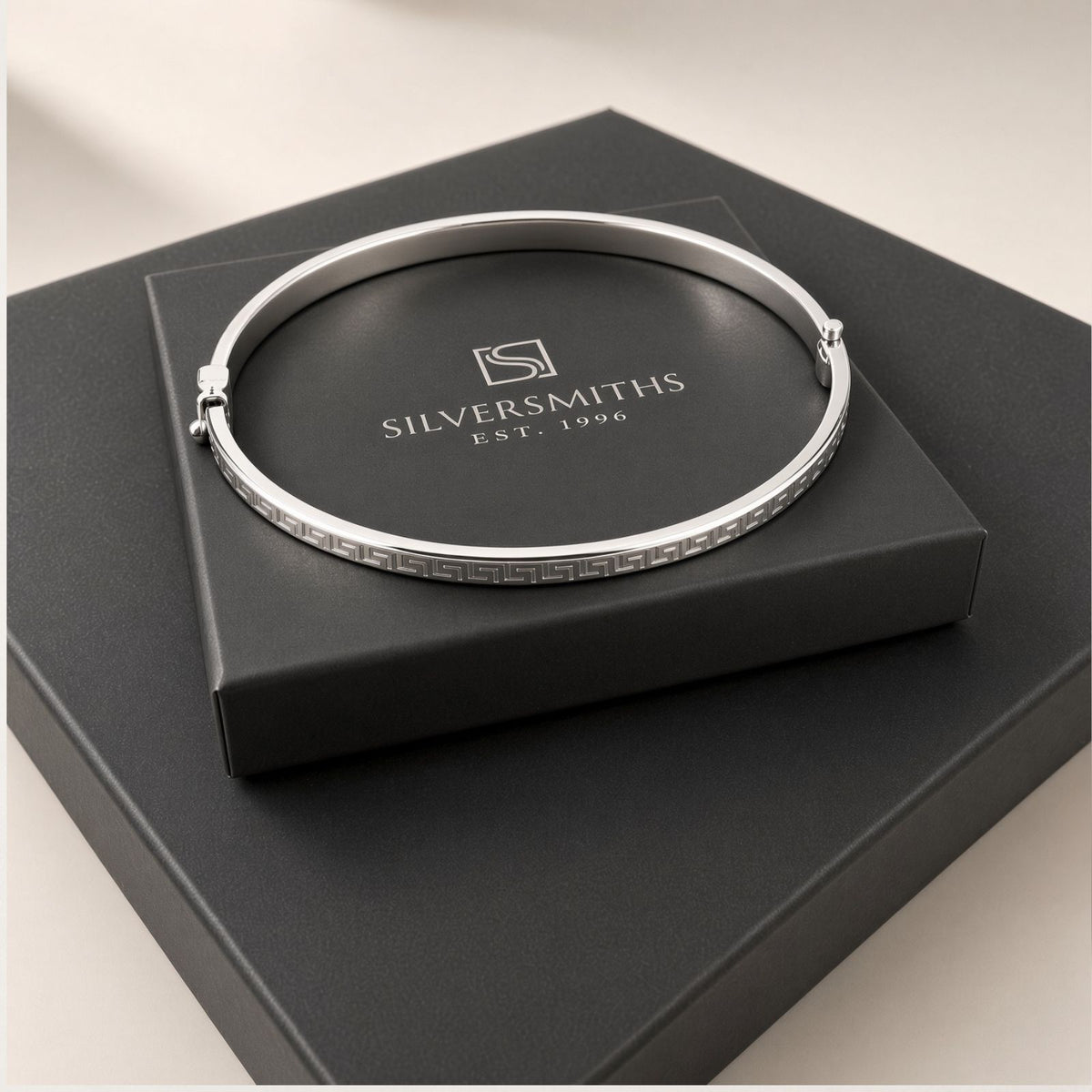 A silver bracelet with a geometric pattern sits on a black Silversmiths box with a white logo and text.