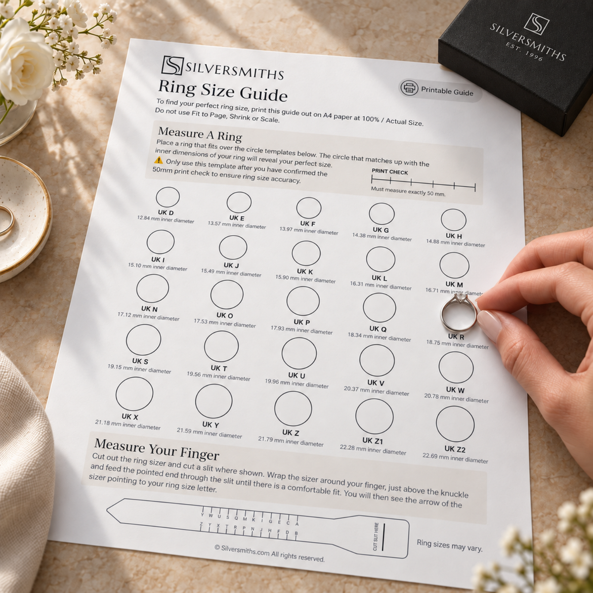 A hand holds a ring above a printed ring size guide on a sunlit table, next to a black jewelry box.