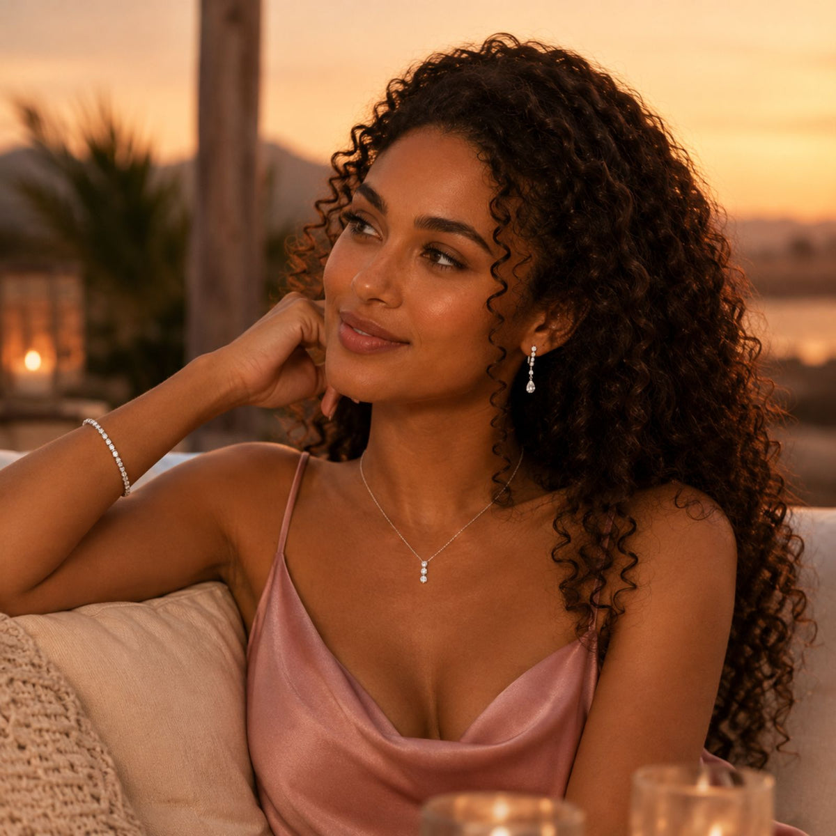 A curly-haired woman in a satin dress and jewelry sits outdoors at sunset, chin resting on her hand.